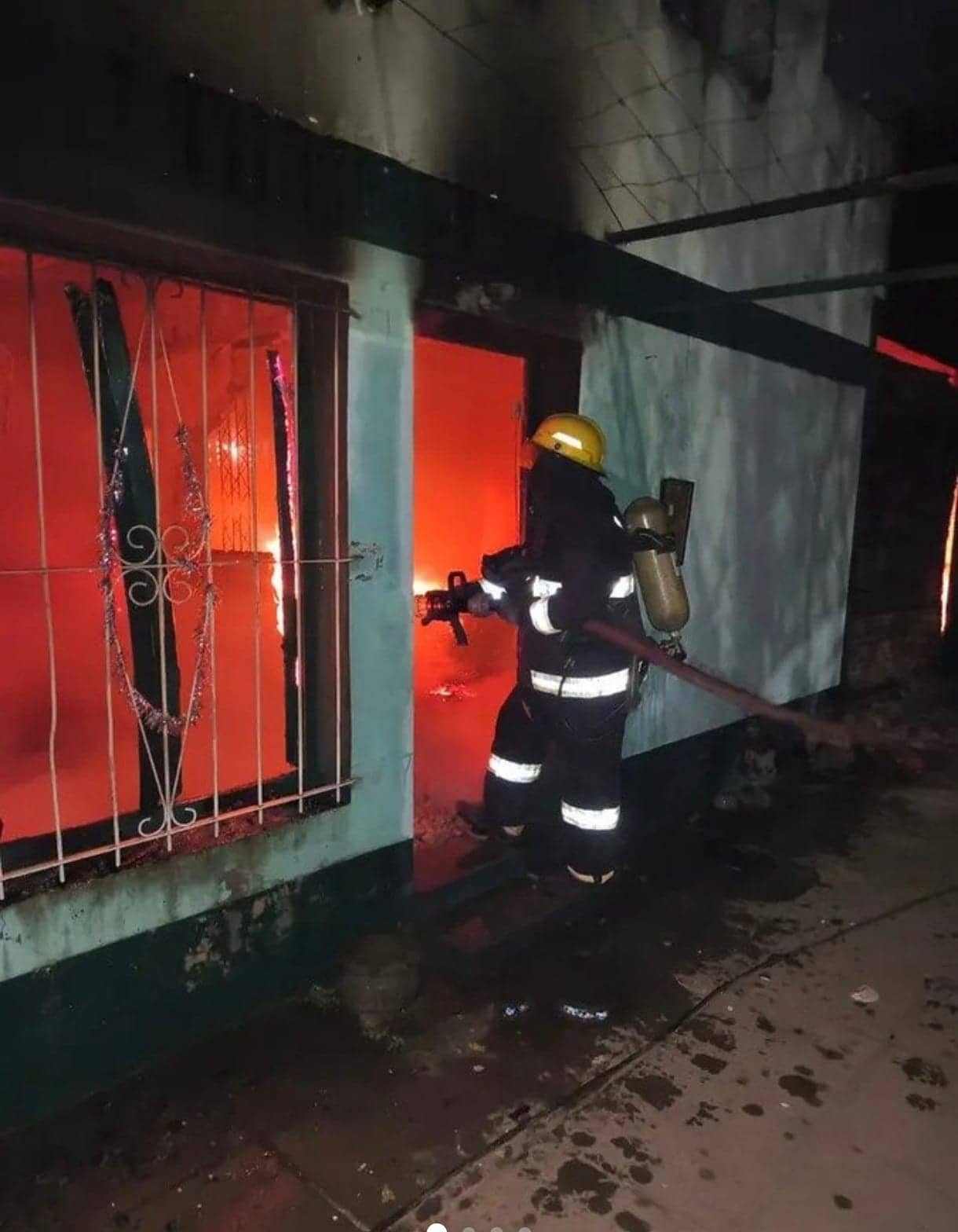 A firefighter in a helmet and breathing gear steps through the door of the house at night. The wall is sea-green; through the barred window the orange glow of the fire is visible inside.