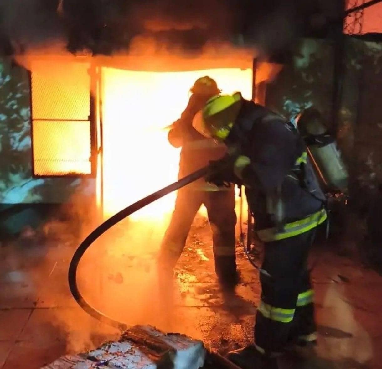 Two firefighters with a hose in front of an opening from which high flames and black smoke are coming. The water has already wet the floor.