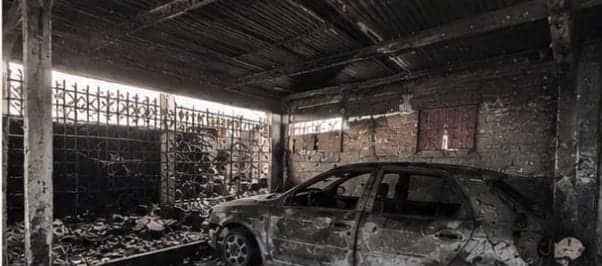 The garage with a burned-out car in the centre. The walls and the sheet-metal roof are black and the floor is covered in burned remains.