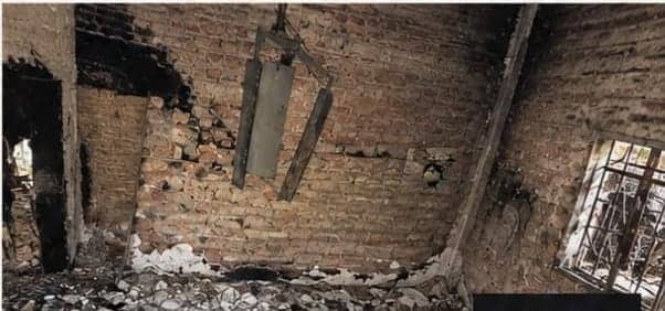 A room with no roof. The walls are bare brick, black in one corner; a burned timber beam hangs in the empty space and the floor is covered in rubble.