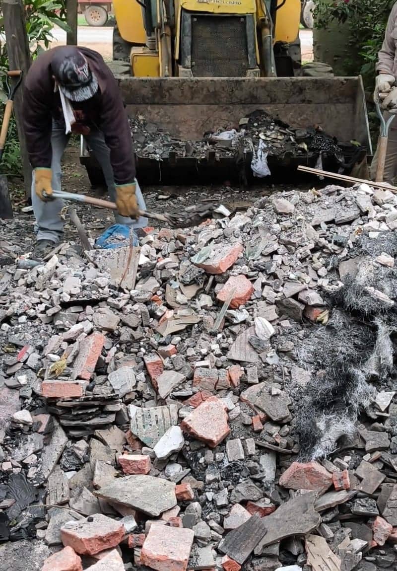 A man with a shovel piles rubble into a mound of broken brick and ash. Behind him, the bucket of a yellow loader is already full of burned remains.