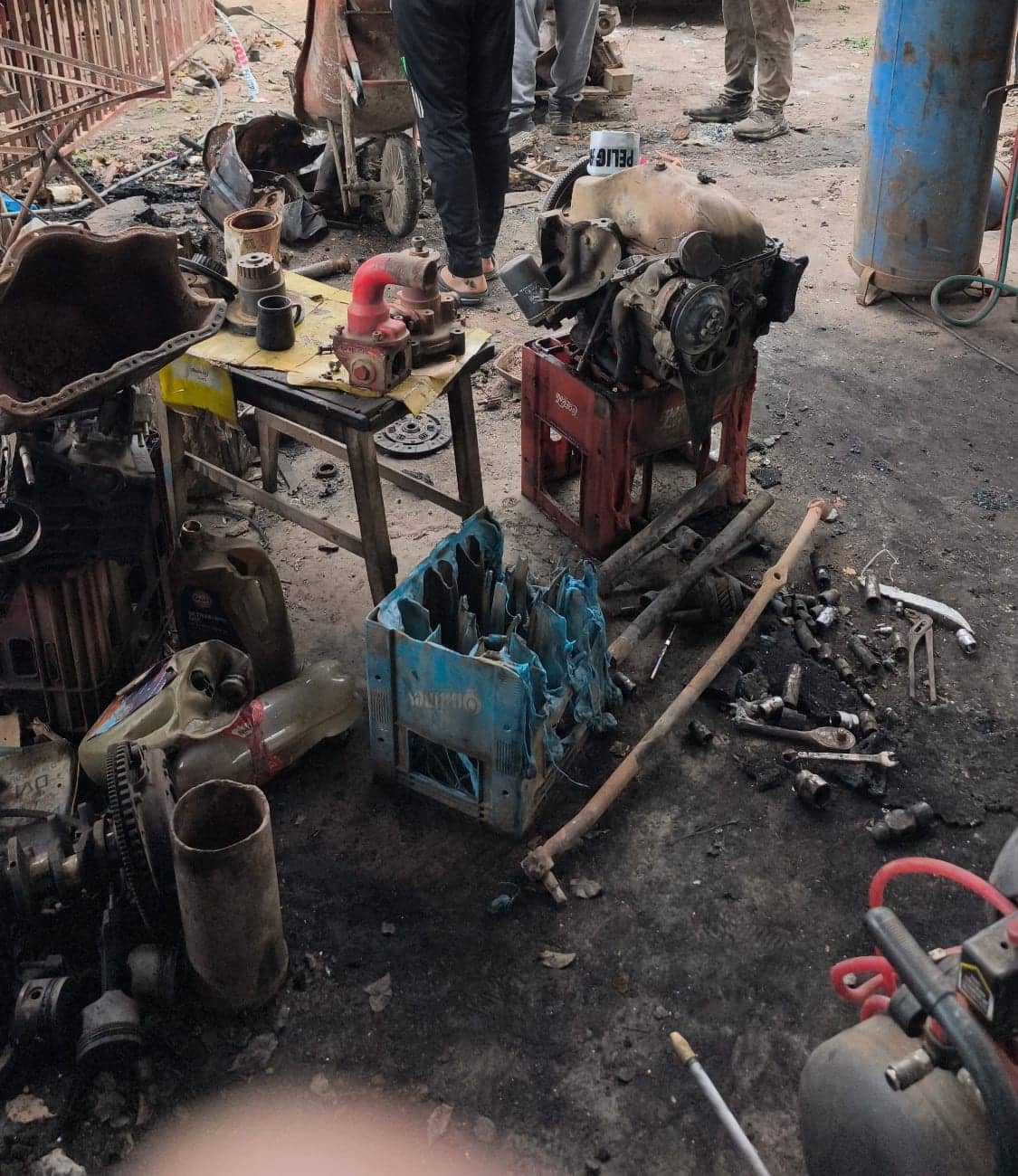 Burned engine parts laid on the floor and on a table, sorted by type: pipes, exhaust manifolds, a blue crate of tools. The legs of several people working around them are visible.