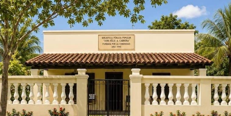 Front of the Don Félix Atilio Cabrera Popular Public Library: a cream building with a tiled roof, a black gate and a plaque on the wall naming the library and the year 1990.