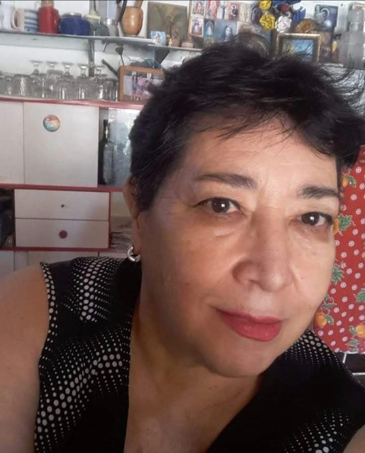 Norma looks at the camera, smiling slightly. She is in her kitchen, wearing a black blouse with white dots. Behind her, a cupboard with glasses and a shelf of framed family photographs.