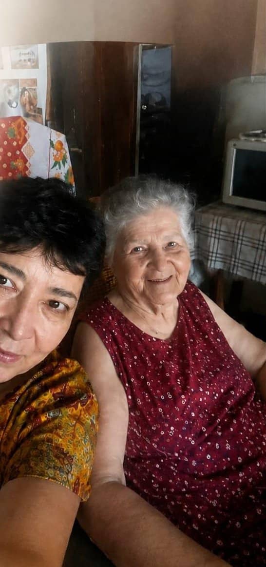 Norma, in a yellow floral blouse, with her mother, Luisa Caballero, who is smiling in a red dress with white dots. They are inside a house.