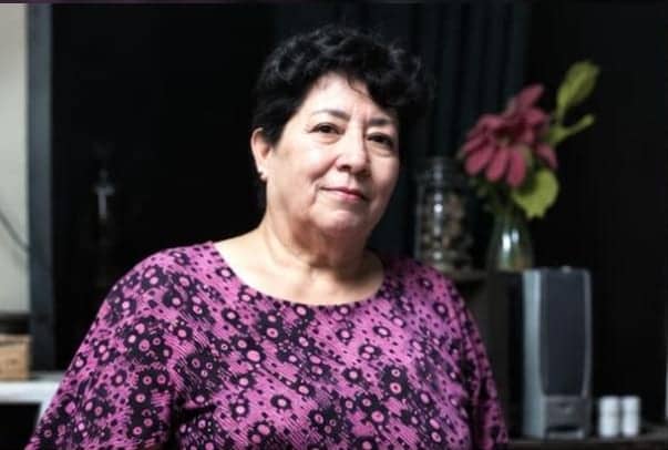 Norma de pie en la cocina, con una blusa fucsia con flores estampadas. A su lado hay un florero con una flor rosa abierta.