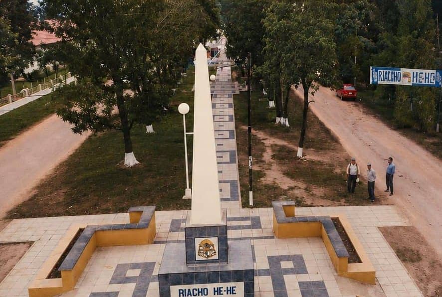 Aerial view of the Riacho He Hé Obelisk. The white monument is seen from above, with the town coat of arms and name on the base, a tiled path, and three men standing to one side.