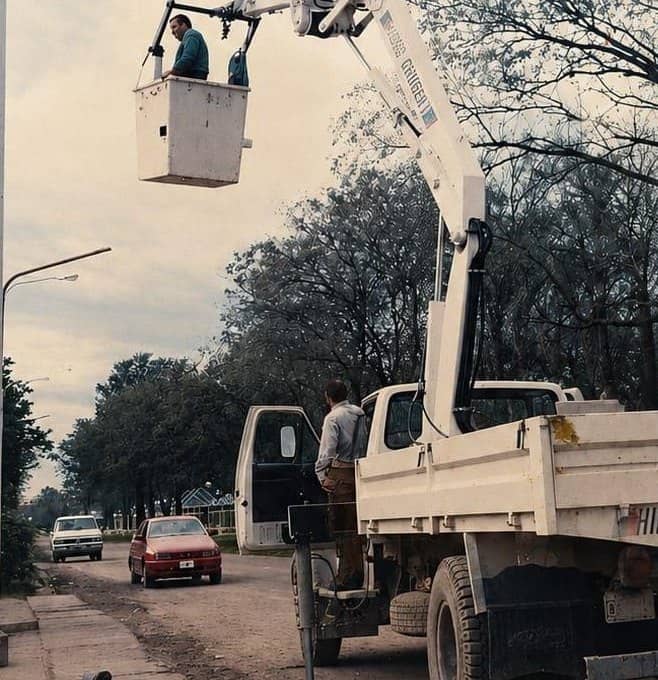 Un hombre en el canasto de un montacargas municipal, elevado sobre la calle. Otro hombre está en el estribo del camión. Al fondo, autos y una hilera de árboles.