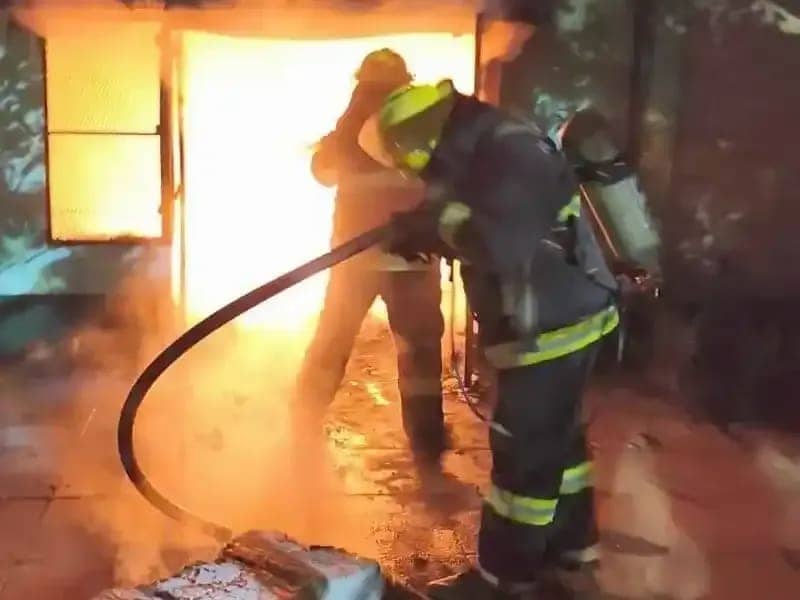 Dos bomberos frente a la puerta de la casa, de noche, con la manguera desplegada contra el fuego.