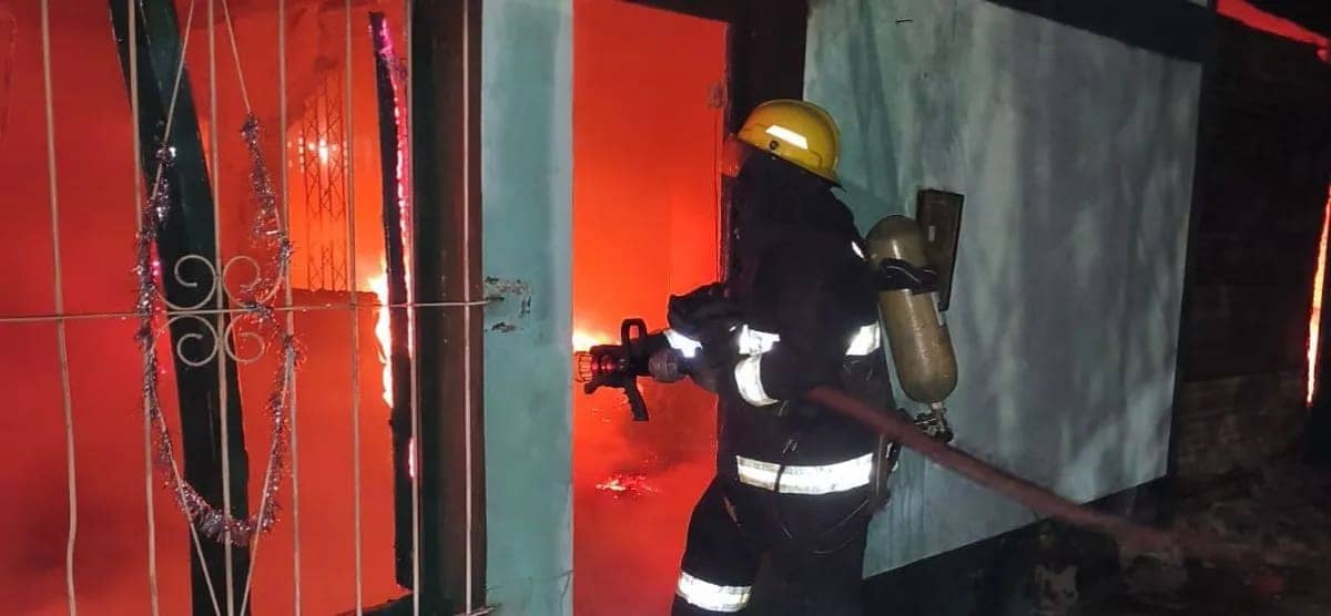 Un bombero con casco amarillo apunta la lanza hacia la puerta, de noche: adentro se ve el resplandor del fuego.