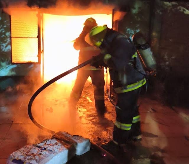 Dos bomberos frente a la puerta de la casa, de noche: uno apunta la manguera y el otro está contra el resplandor del fuego.